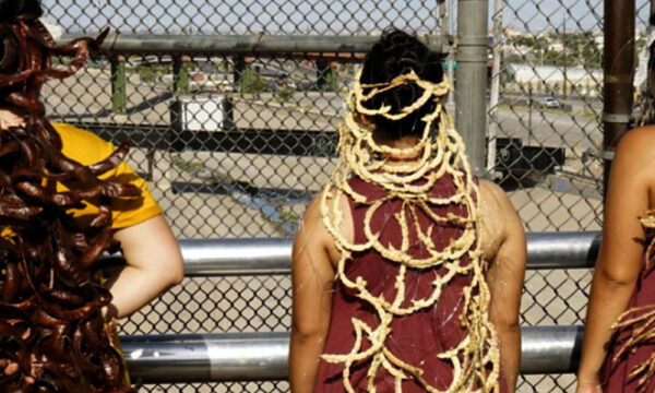 Three women performing artists stand on a bridge between El Paso and Juarez, with a crown of seeds cascading down their backs