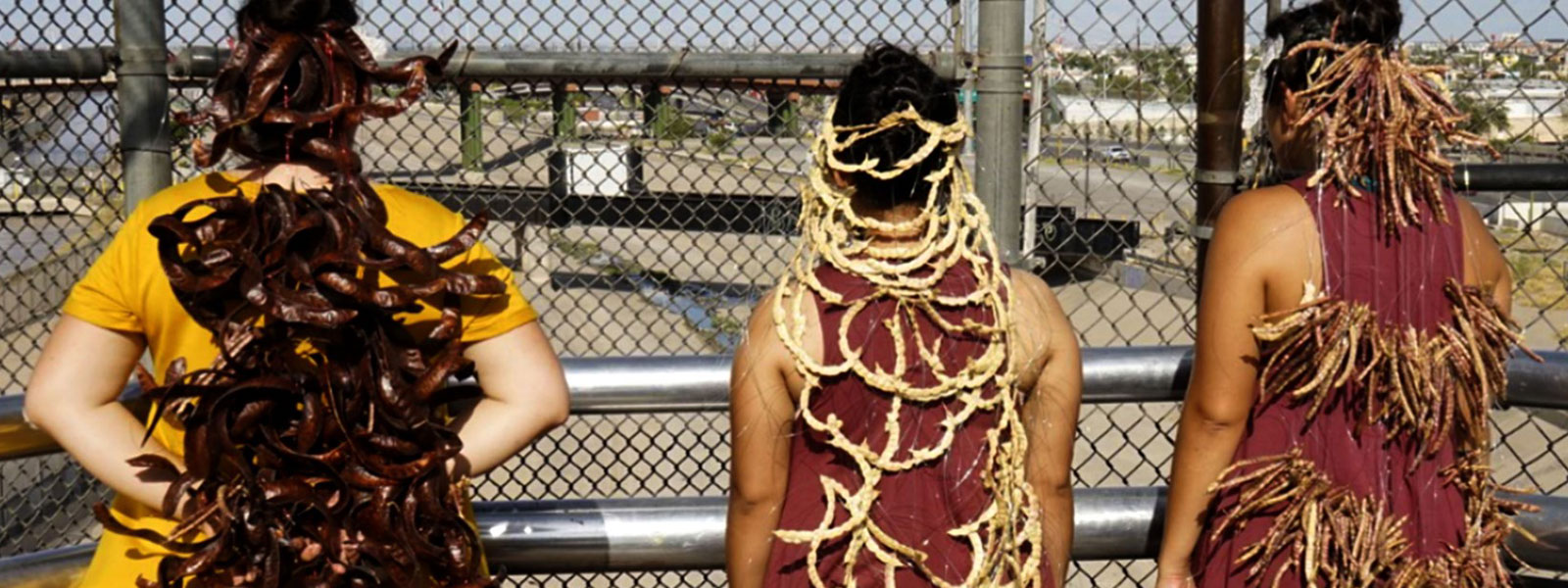 Three women performing artists stand on a bridge between El Paso and Juarez, with a crown of seeds cascading down their backs