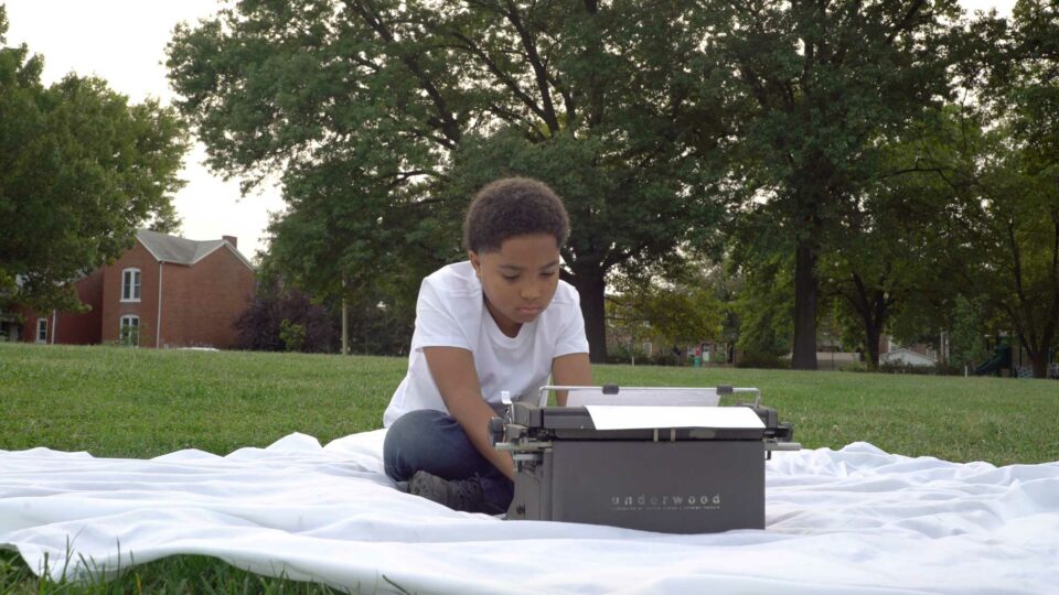 film still of a young black boy, typing on a typewriter on a blanket in the middle of a grass field