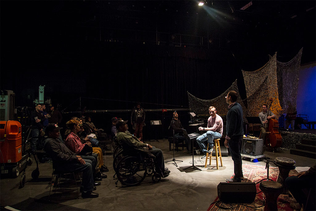 man onstage, seated on a stool in a spotlight