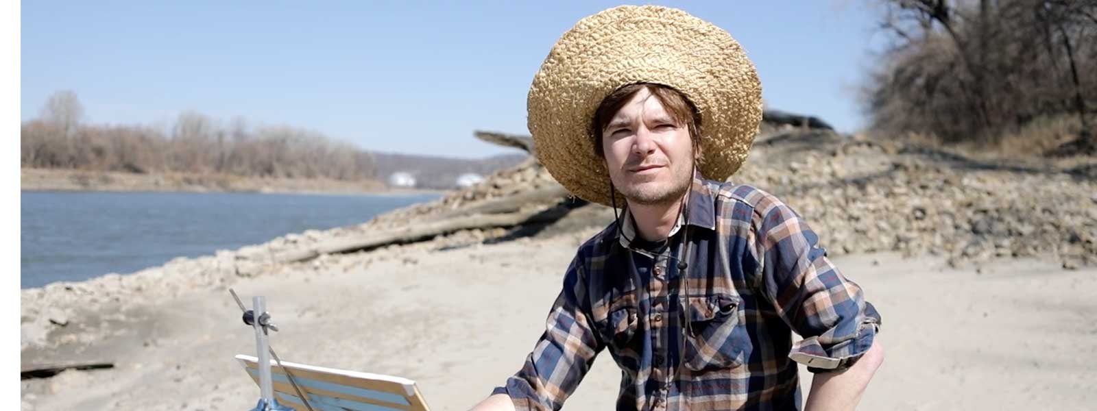 man in hat sits at easel by river