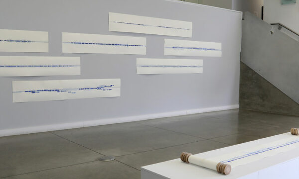 gallery installation of horizontal works of paper hung on wall, with a work on paper on scroll on a plinth