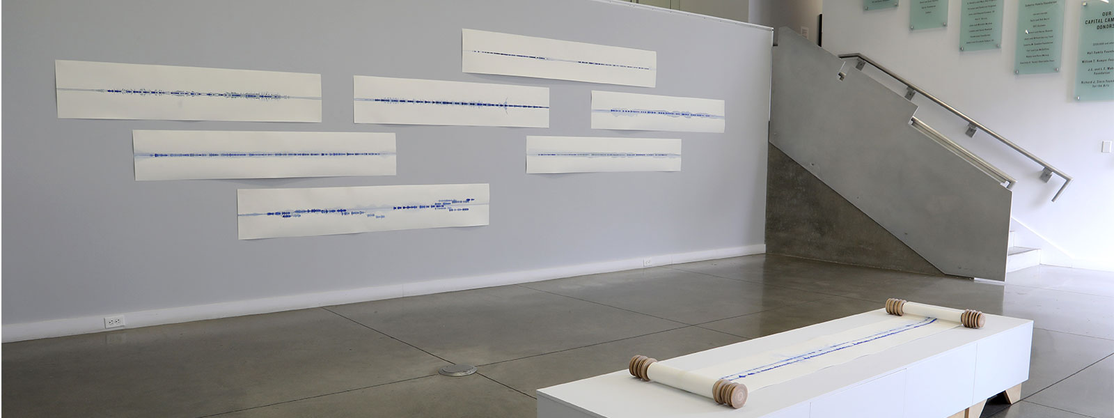 gallery installation of horizontal works of paper hung on wall, with a work on paper on scroll on a plinth