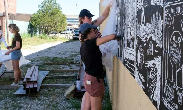 Two women on the right of the image hang prints on the side of a beige building