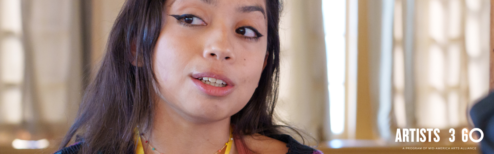 Woman with dark hair and dark eyeliner looks to the left and speaks.