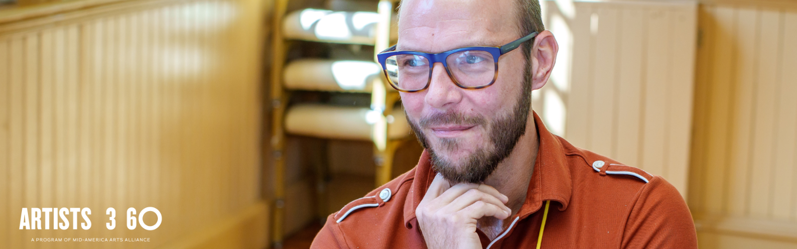 Man wearing glasses, a yellow lanyard around his neck, and dark orange shirt holds his hand up to his face in a thinking pose.