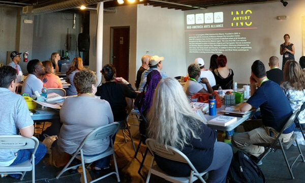 People sit in an art gallery at folding tables watching a presentation by an artist.