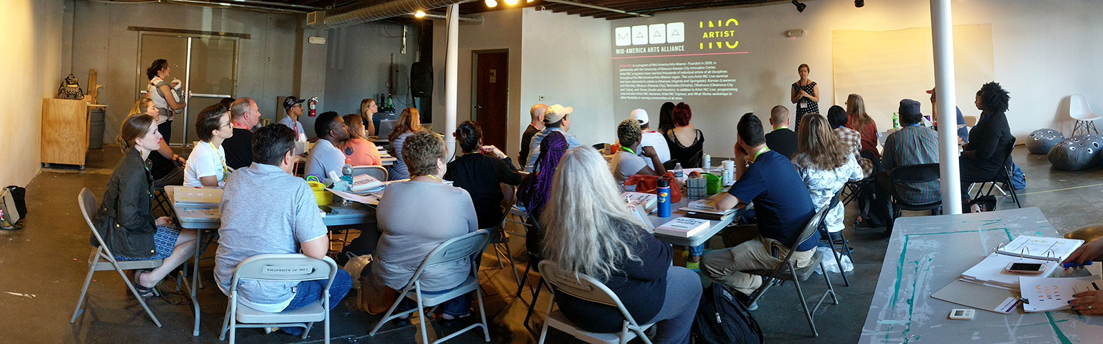 People sit in an art gallery at folding tables watching a presentation by an artist.