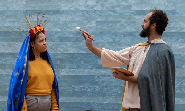 A woman wears a gold shirt, cape, and crown, while a man holds a paintbrush in a theatrical production.