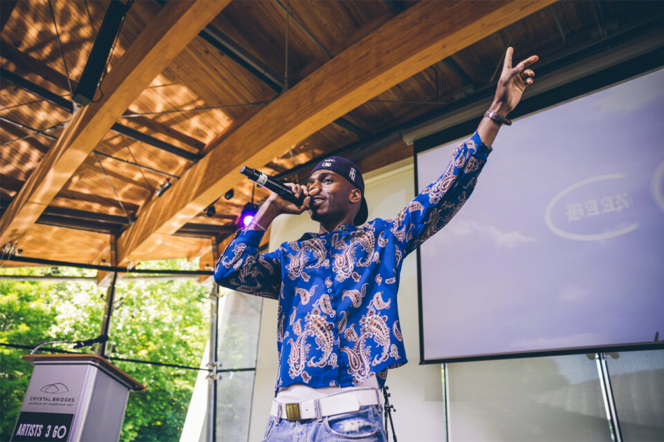 A young man with a blue patterned shirt holds a microphone to his mouth speaking on a stage with an enthusiastic arm in the air.