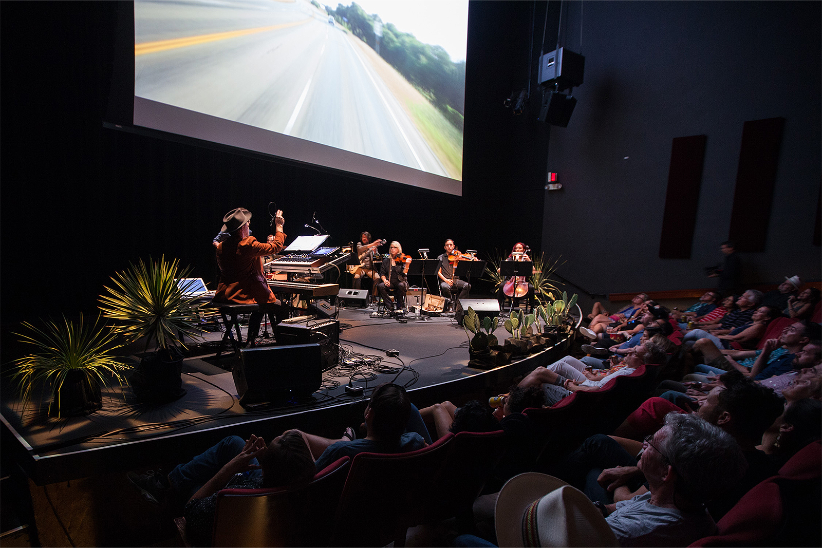 People sit in an audience looking a stage with a visual projection and musicians playing instruments.