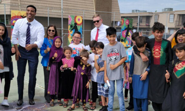 Children, staff, and teachers stand outside of a building with colorful paintings installed on a fence.