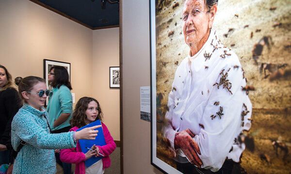 Woman and girl look at framed photograph in gallery of a woman with bees on her clothes.