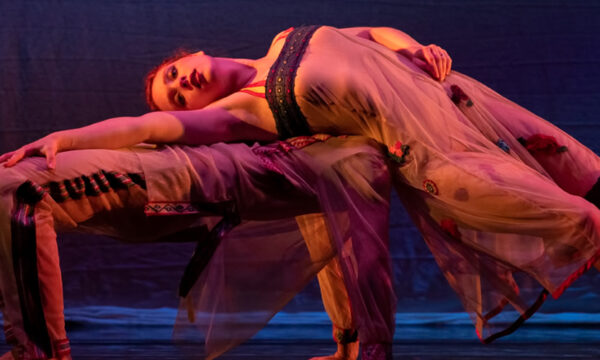 A graceful female dancer in silky dress arches over a male dancer also arching with dramatic warm lighting and cool blue light in the background.