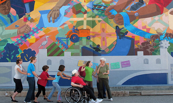 A small group of people, from young to old, with one person in red shirt in a wheelchair, dance together along a huge colorful outdoor mural.