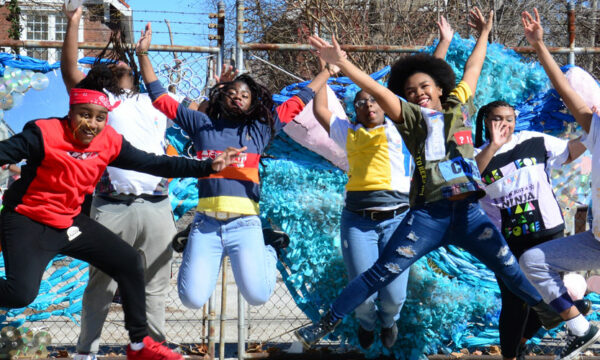 People jump up in the air with smiles on faces and a bright blue mural painting in background.