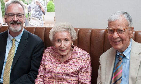 Man with suit on left smiles with two elderly individuals: one woman with red suit, a man with tan suit.