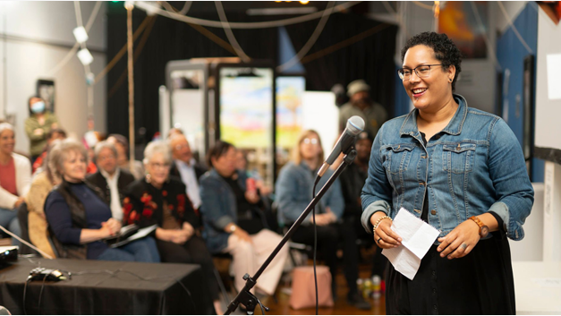 Black woman artists stands in front of a microphone presenting her art to a crowd of Artist INC alumni