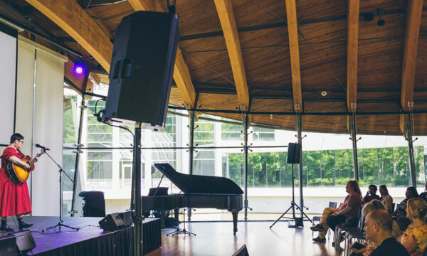 Woman on left in red dress holds guitar while singing onstage with audience on the right in large auditorium with wooden beams on ceiling.