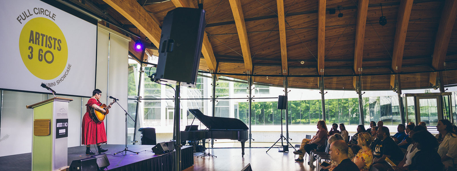 Woman on left in red dress holds guitar while singing onstage with audience on the right in large auditorium with wooden beams on ceiling.