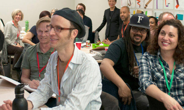 Artists sit in a classroom with books and notebooks on their shared tables. People are smiling turned towards the camera.