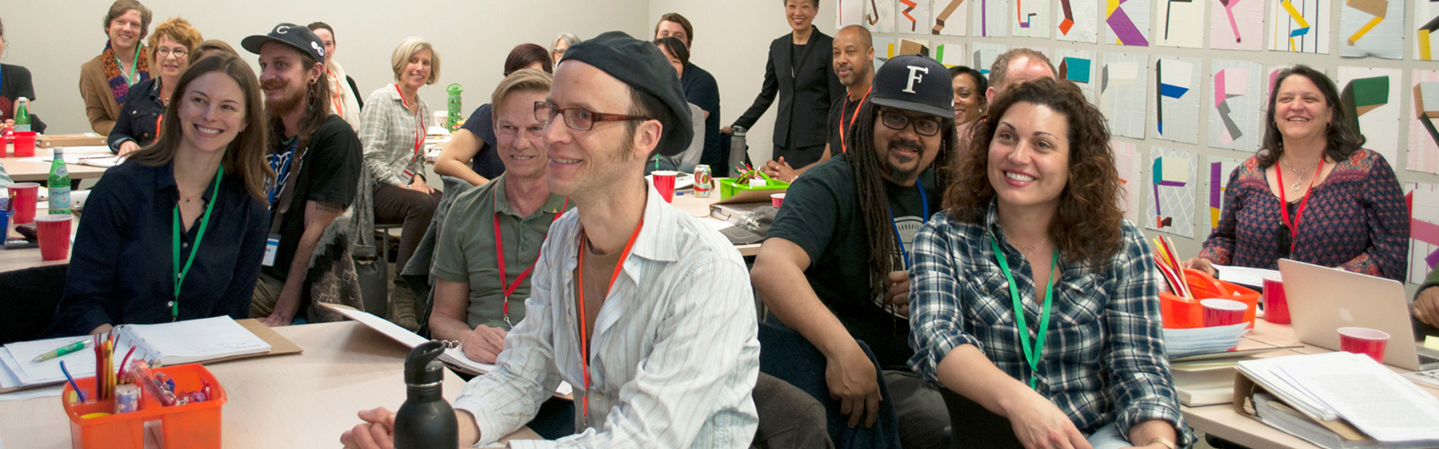 Artists sit in a classroom with books and notebooks on their shared tables. People are smiling turned towards the camera.