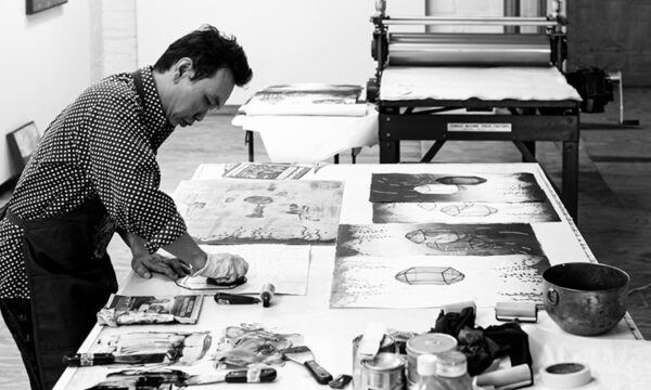 Male artist leans over a work table creating a print in a printmaking studio with art and materials surrounding him.