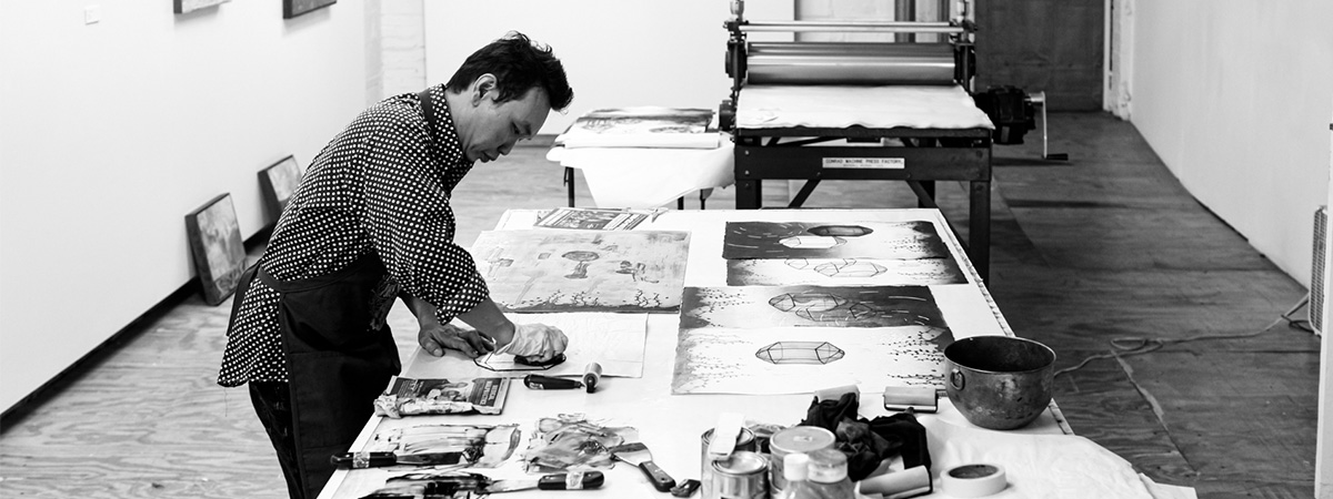 Male artist leans over a work table creating a print in a printmaking studio with art and materials surrounding him.