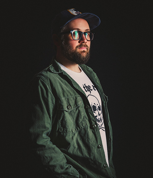 A man wears a casual green over shirt and white t-shirt with glasses and a baseball hat looks at the camera for a headshot with a shadowy black background. 