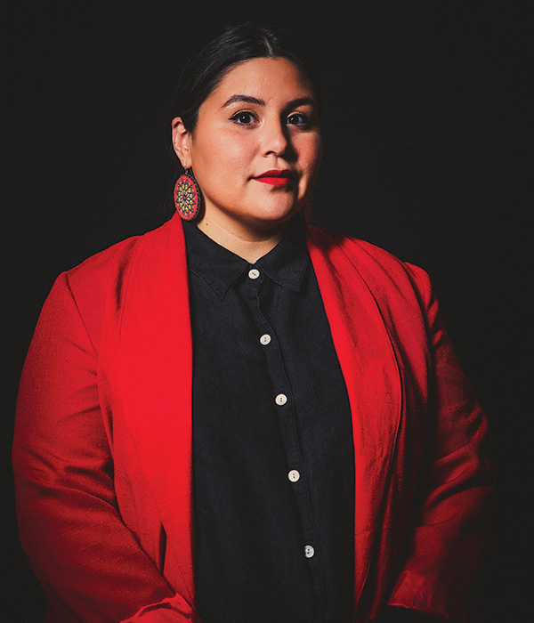 A person with a red blazer, black button down shirt, large golden earrings, and black hair with red lipstick looks at camera for a posed headshot with a shadowy black background