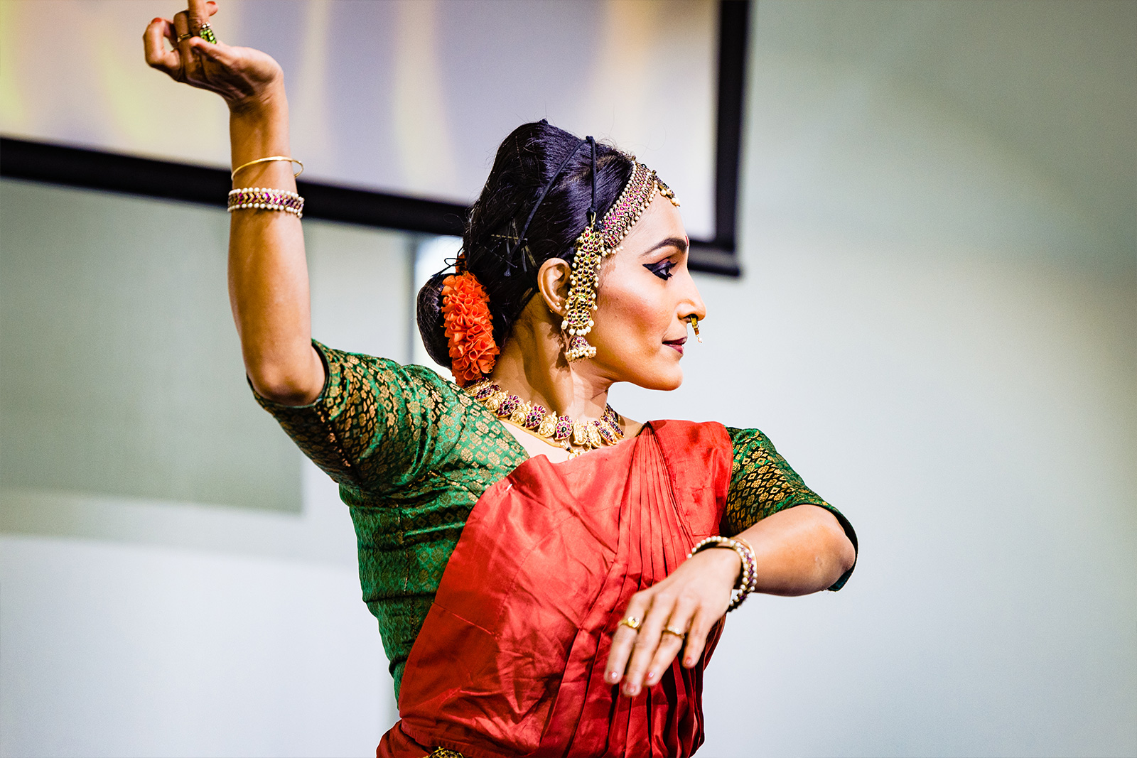 Woman wearing red and green clothing has her arm held high and fingers raised and curved in different shapes as she looks over her shoulder in movement.