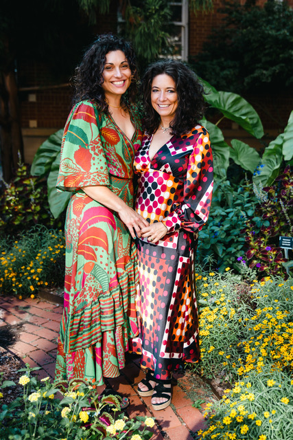 Two women in bright colorful dresses stand together holding hands with a garden in the background.