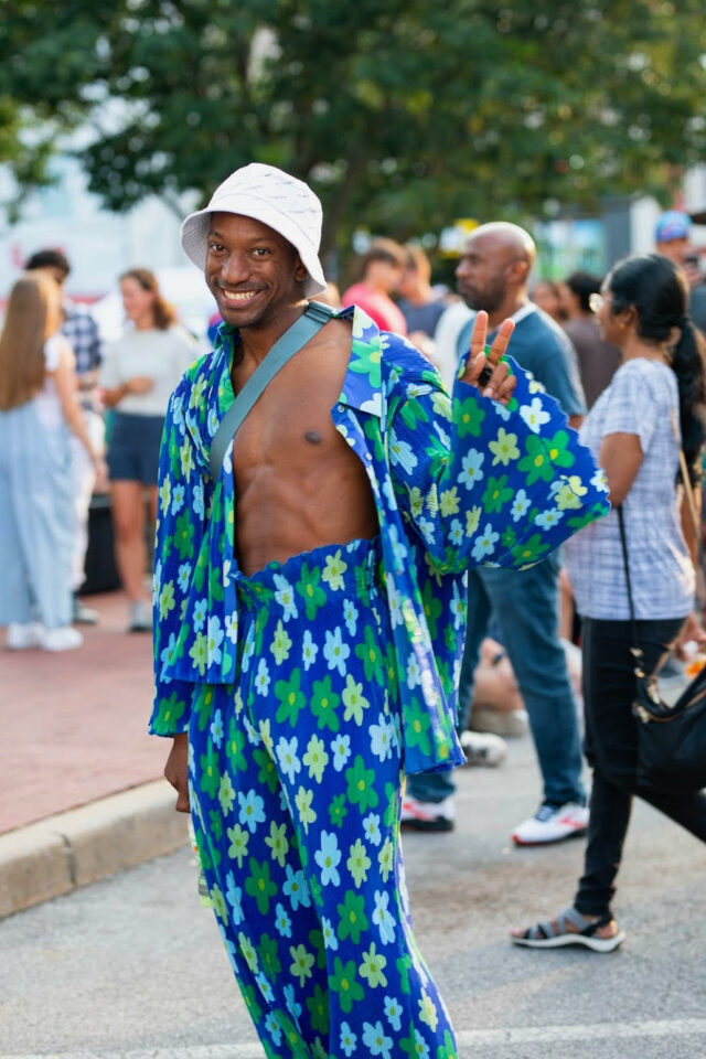 Man with white hat wearing shirt and pants with a pattern of blue flowers looks at camera with a smile.