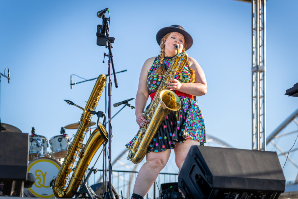 Person in a dress on an outdoor stage plays saxophone.