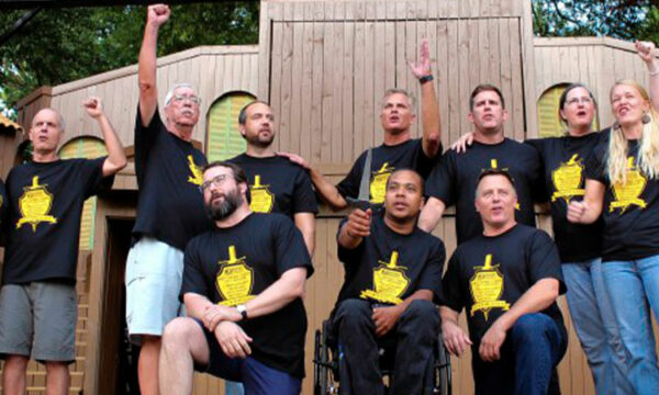 A group of performers on a stage. While most people are standing with their hands or first in the air, two men kneel beside a performer in a wheelchair.