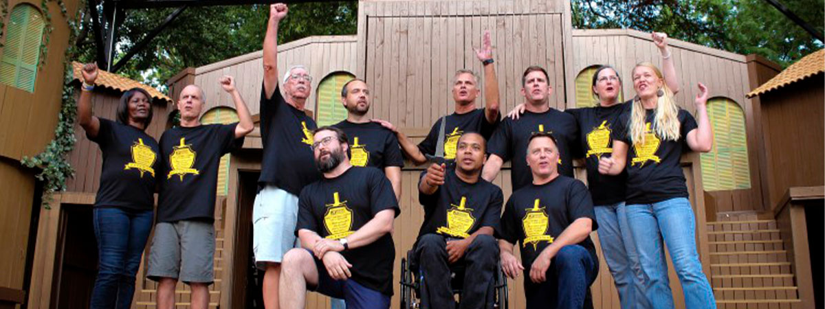 A group of performers on a stage. While most people are standing with their hands or first in the air, two men kneel beside a performer in a wheelchair.