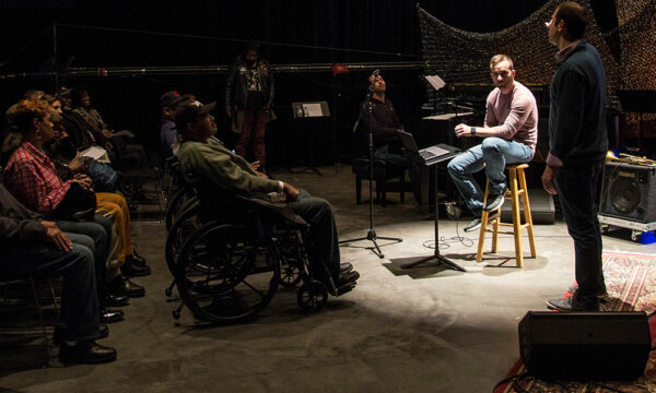 Man sits on a stool with a overhead spotlight with an audience.