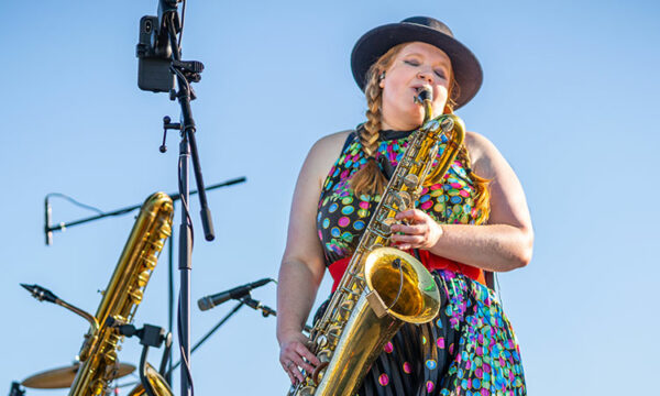 Musician in colorful clothes plays a saxophone on an outdoor stage with a blue sky in the background.