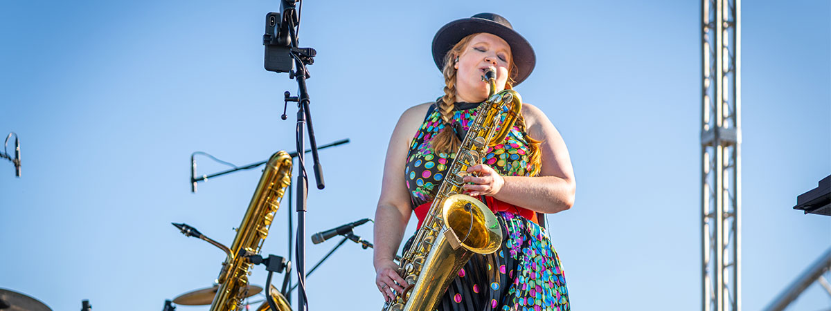 Musician in colorful clothes plays a saxophone on an outdoor stage with a blue sky in the background.