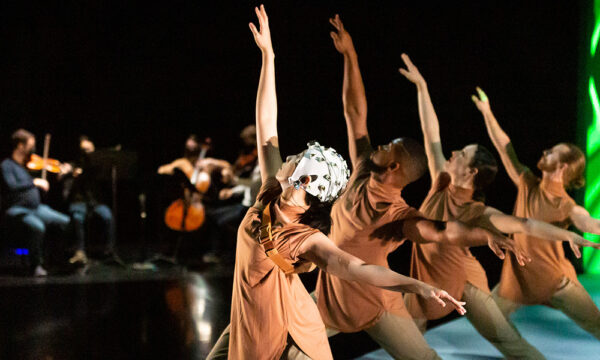 Four dancers step out in a lunge with their right arm high. Dancers wear a cap on their heads and tan costumes. There are bright neon lights onstage in the background that appear to be vertical waves. There are also musicians playing string instruments in the background on the left.