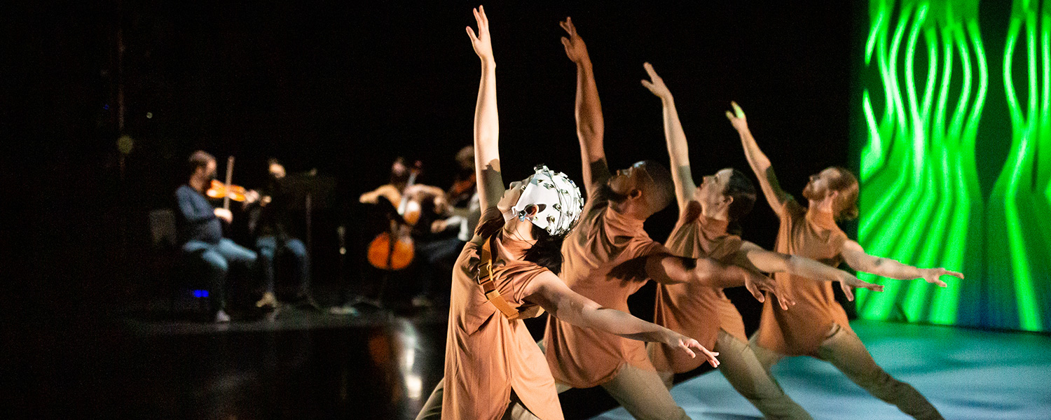 Four dancers step out in a lunge with their right arm high. Dancers wear a cap on their heads and tan costumes. There are bright neon lights onstage in the background that appear to be vertical waves. There are also musicians playing string instruments in the background on the left.