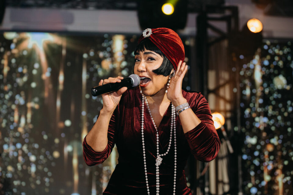 Woman wearing jazzy red hat and dress with long necklaces sings into a microphone with a background of shiny, reflective decor.