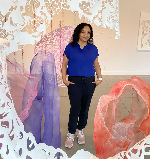 Artist Sukanya Mani wears a blue shirt and black pants standing in her installation of cut paper and painted wire mesh. The paper and mesh surround her, framing her in the gallery.