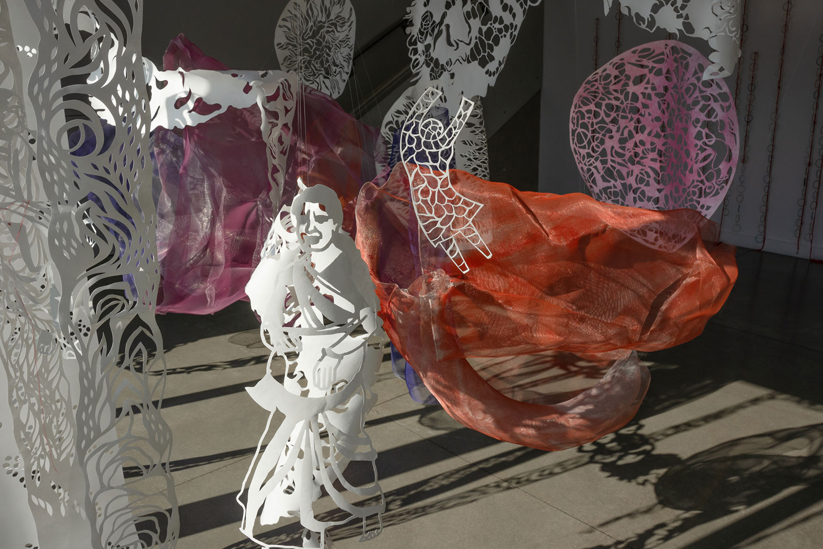 Paper cuttings of a woman and a child float hanging in an art gallery with pink and orange wire mesh in the background.
