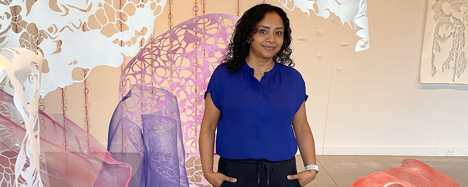 Artist Sukanya Mani in a blue shirt standing in her cut-paper installation at the M-AAA Culture Lab gallery.