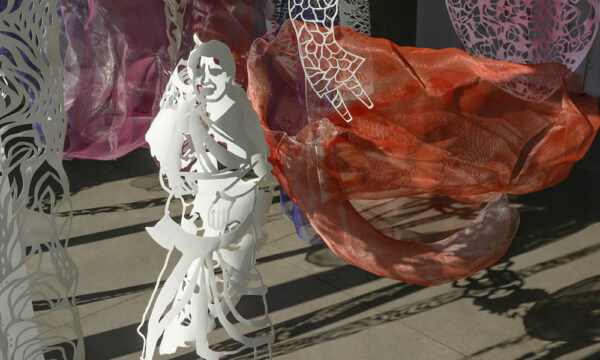 Cut paper detail featuring a woman with a background of a gallery with hanging orange and pink wire mesh sculptures.