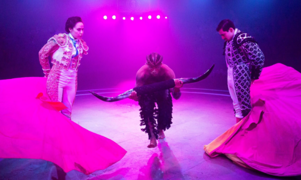 Two matadors with capes in bright pink stage lighting swing the capes as a person with bull horns runs through the middle.