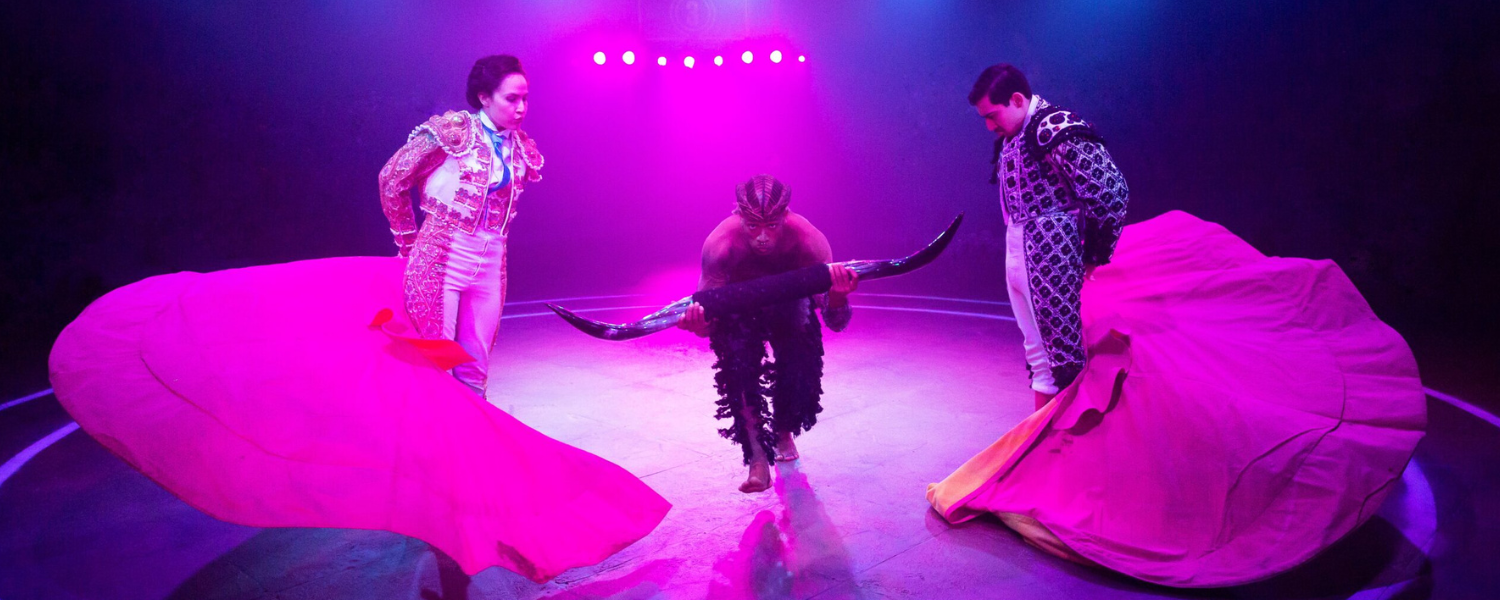 Two matadors with capes in bright pink stage lighting swing the capes as a person with bull horns runs through the middle.