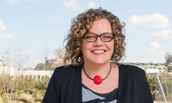 Woman with a black cardigan and black and white patterned shirt, necklace with red pendant, and with curly hair and glass smiles standing in front of a cityscape.