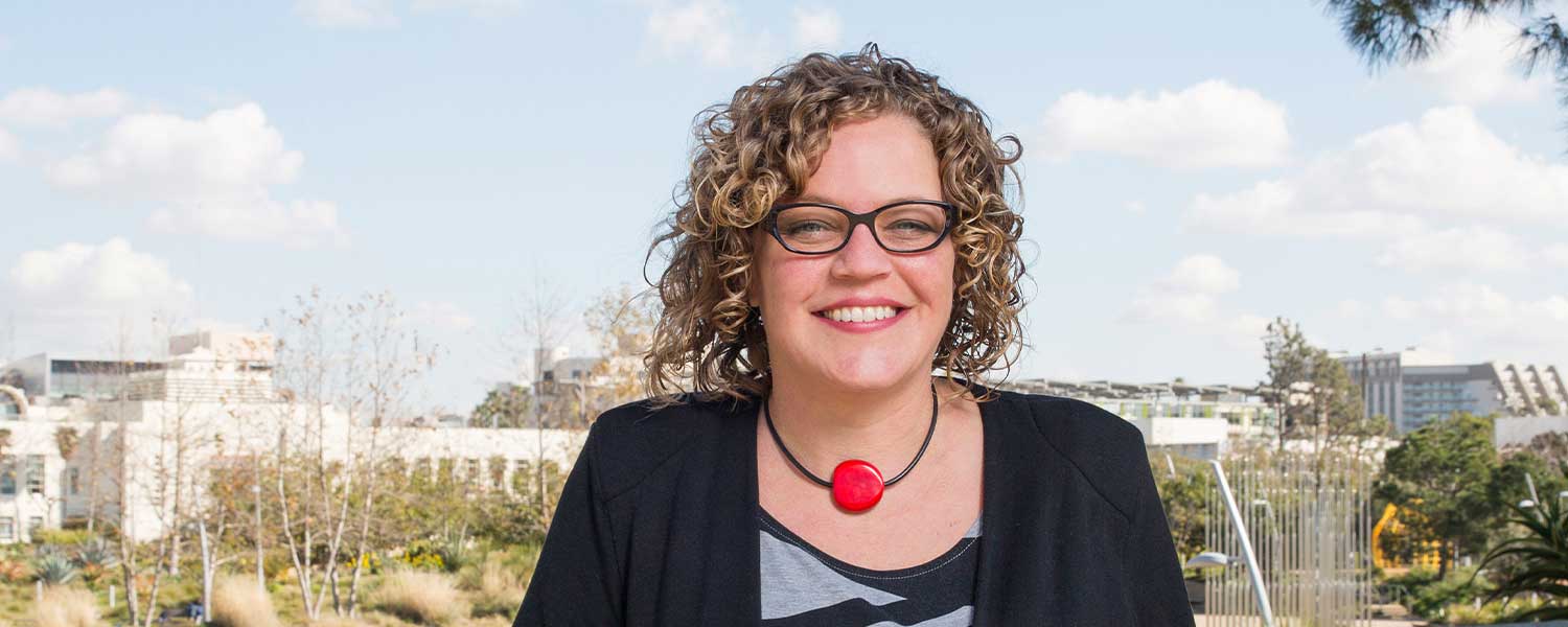 Woman with a black cardigan and black and white patterned shirt, necklace with red pendant, and with curly hair and glass smiles standing in front of a cityscape.